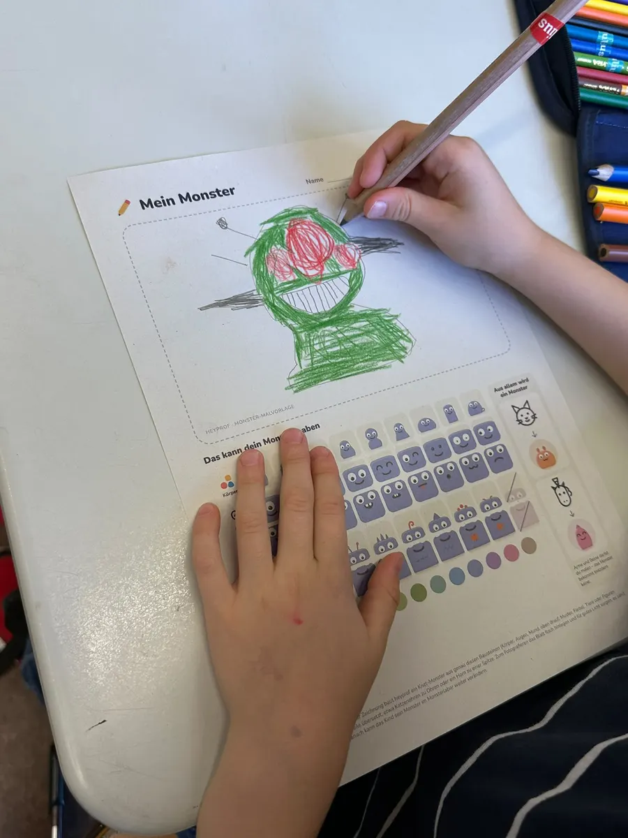Kinderhand malt auf der Malvorlage „Mein Monster“ ein grünes Monster mit drei roten Augen
